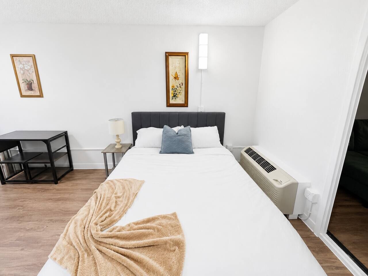 Ett hotellrum med en säng, ett sängbord, en lampa, konst på väggen och en värmare.