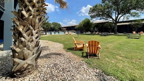 A grassy outdoor area with a palm tree, two Adirondack chairs, and a building in the background.
