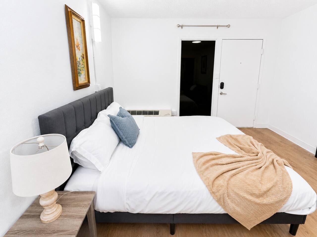 A bedroom with a bed, a nightstand, a lamp, a door, and a framed picture on the wall.