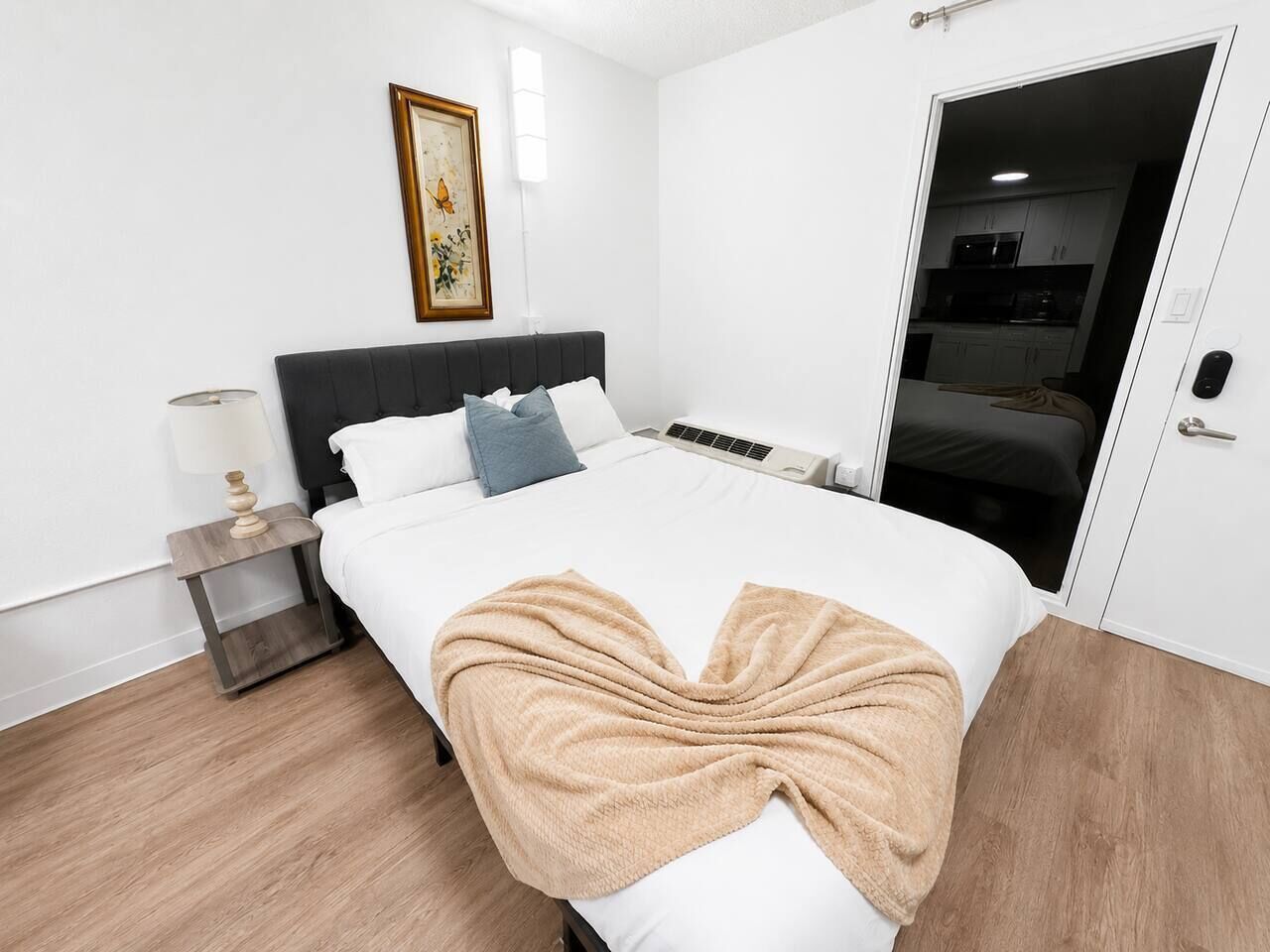 A modern hotel room with a bed, bedside table, lamp, and a door leading to a kitchenette.