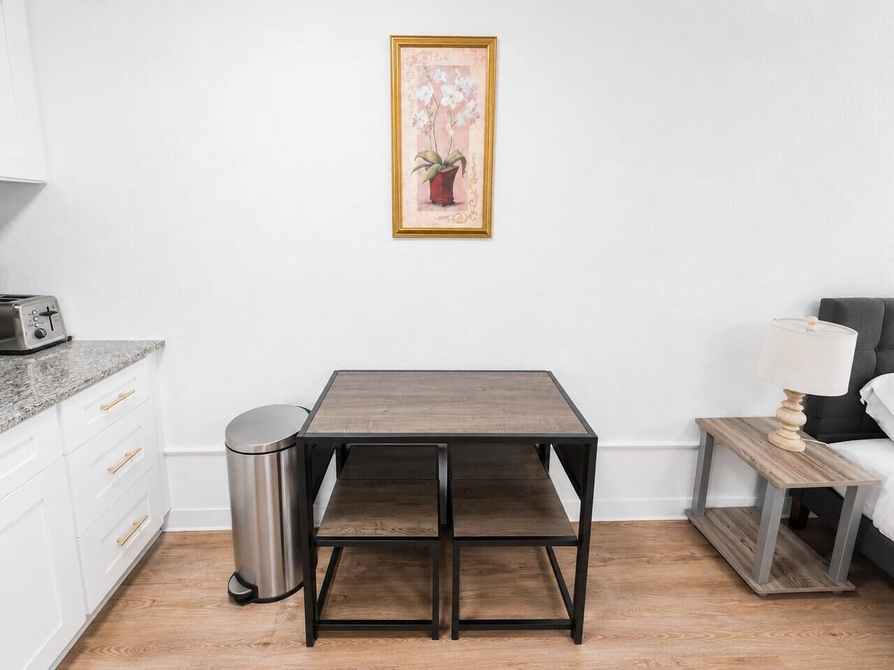 A small, modern kitchen with a wooden table, a trash bin, and a framed picture on the wall.