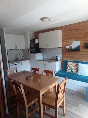 A compact kitchen with white cabinets, a microwave, and a coffee machine. There's a dining table with chairs and a sofa with a colorful pillow. - Apartment for 4–6 people – 100 m from the CENTER STATION – ESF – at the base of the slopes (Valmeinier)