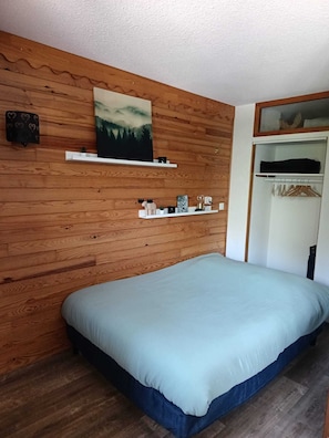 Ein Schlafzimmer mit Holzwand, einem Bett mit hellblau gestreuter Bettdecke, einem Regal mit Dekoration und einem Kleiderschrank mit Holzaufhängern.