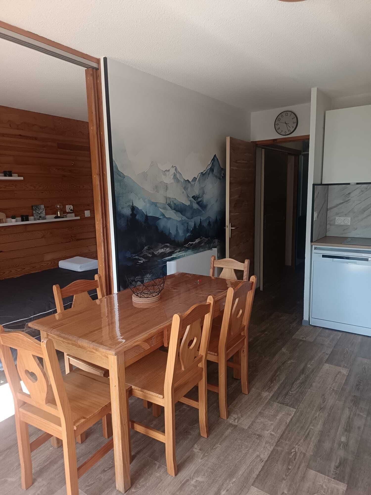 A dining area with a wooden table and chairs, a kitchen with a refrigerator, and a wall with a mountain landscape mural.