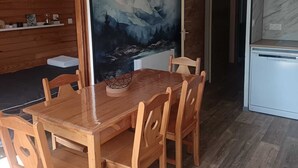 Et spiseområde med et træbord og stole, et køkken med et køleskab, og en væg med et bjerglandskabsmaleri.