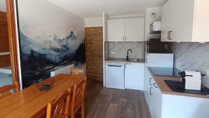 Et kompakt køkken med hvide skabe, et træspisebord og et vægmaleri af et bjerglandskab.