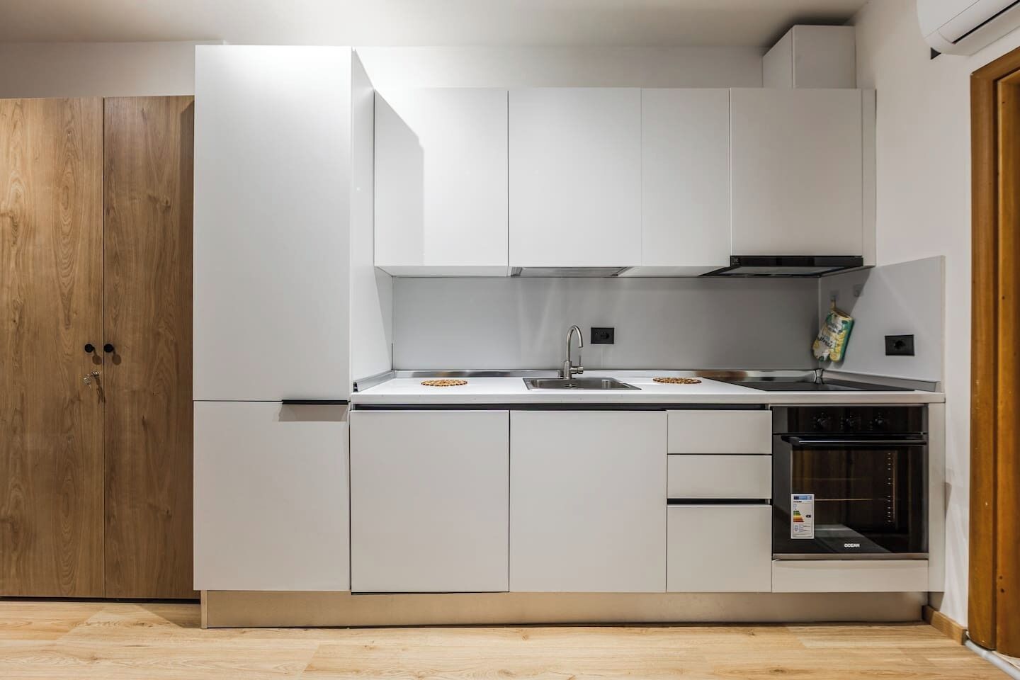 A modern kitchen with white cabinetry, a wooden door, a sink, and an oven.