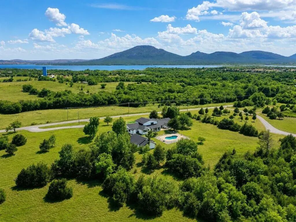Ett bostadsområde med ett hus omgivet av frodig grönska, en swimmingpool och utsikt över en sjö och berg i bakgrunden.