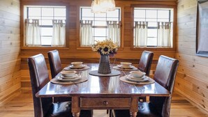 Uma sala de jantar com painéis de madeira, mesa posta para seis pessoas, um vaso de flores e cortinas nas janelas.