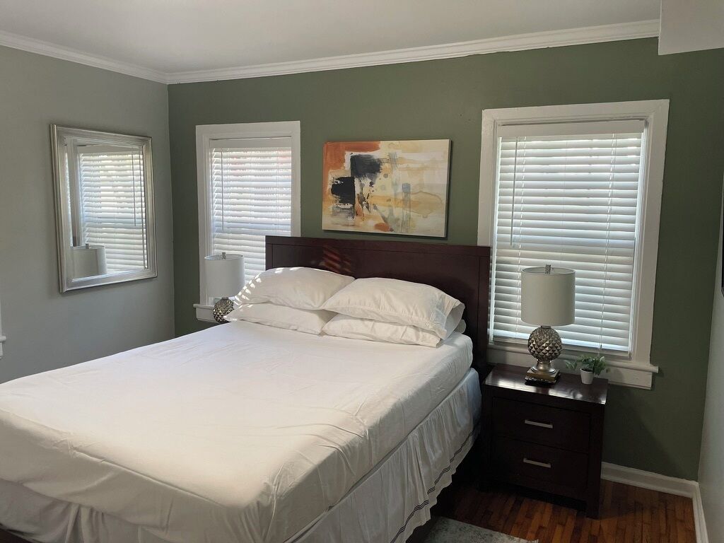 Quarto com cama, mesas de cabeceira, abajur, pintura e janelas com persianas.