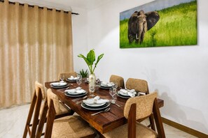A dining area with a wooden table set for six, surrounded by cushioned chairs. A large painting of elephants is on the wall.