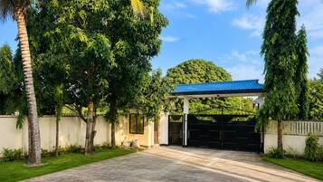 A gated driveway leading to a building with a blue roof, surrounded by lush greenery and a clear sky.