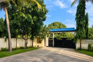 A gated driveway leading to a building with a blue roof, surrounded by lush greenery and a clear sky.