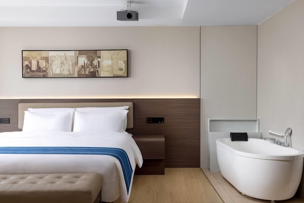 A modern hotel room with a bed, a large artwork on the wall, a side table, a built-in bathtub, and a ceiling-mounted projector.