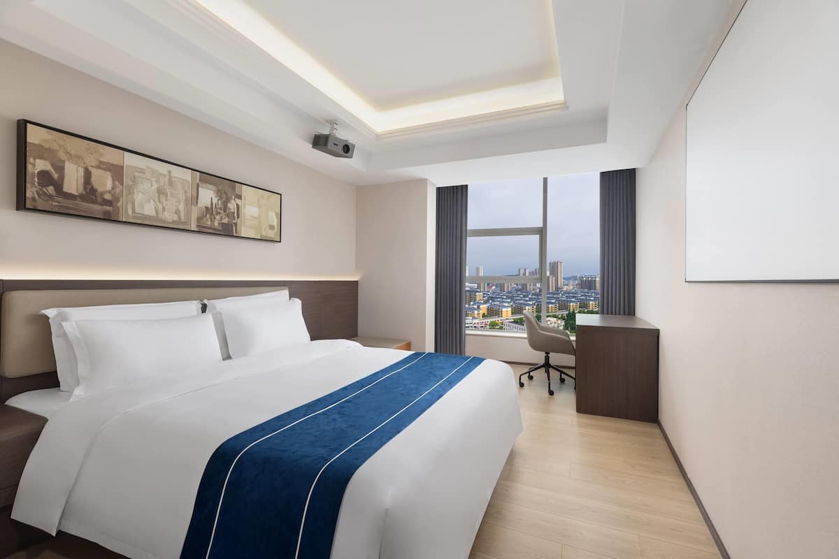 A modern hotel room with a large bed, a desk, a chair, and a city view.