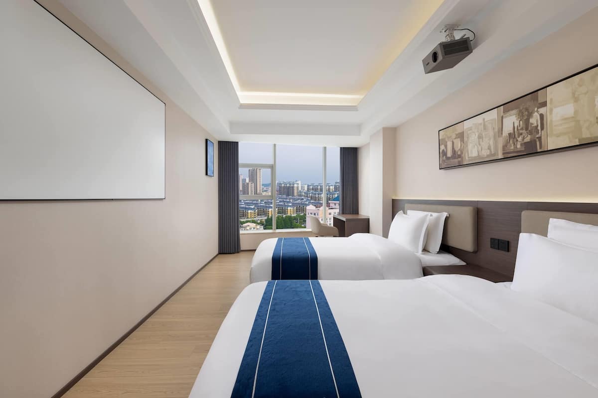 A hotel room with two beds, a city view, a projector, and a framed artwork on the wall.