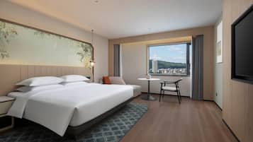 A hotel room with a large bed, a desk, a chair, a TV, and a window with a city view.