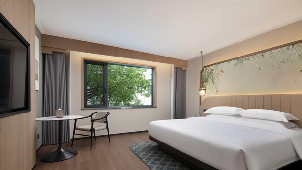 A hotel room with a large bed, a small table with a chair, a window with a view of trees, and a wall with a floral mural.