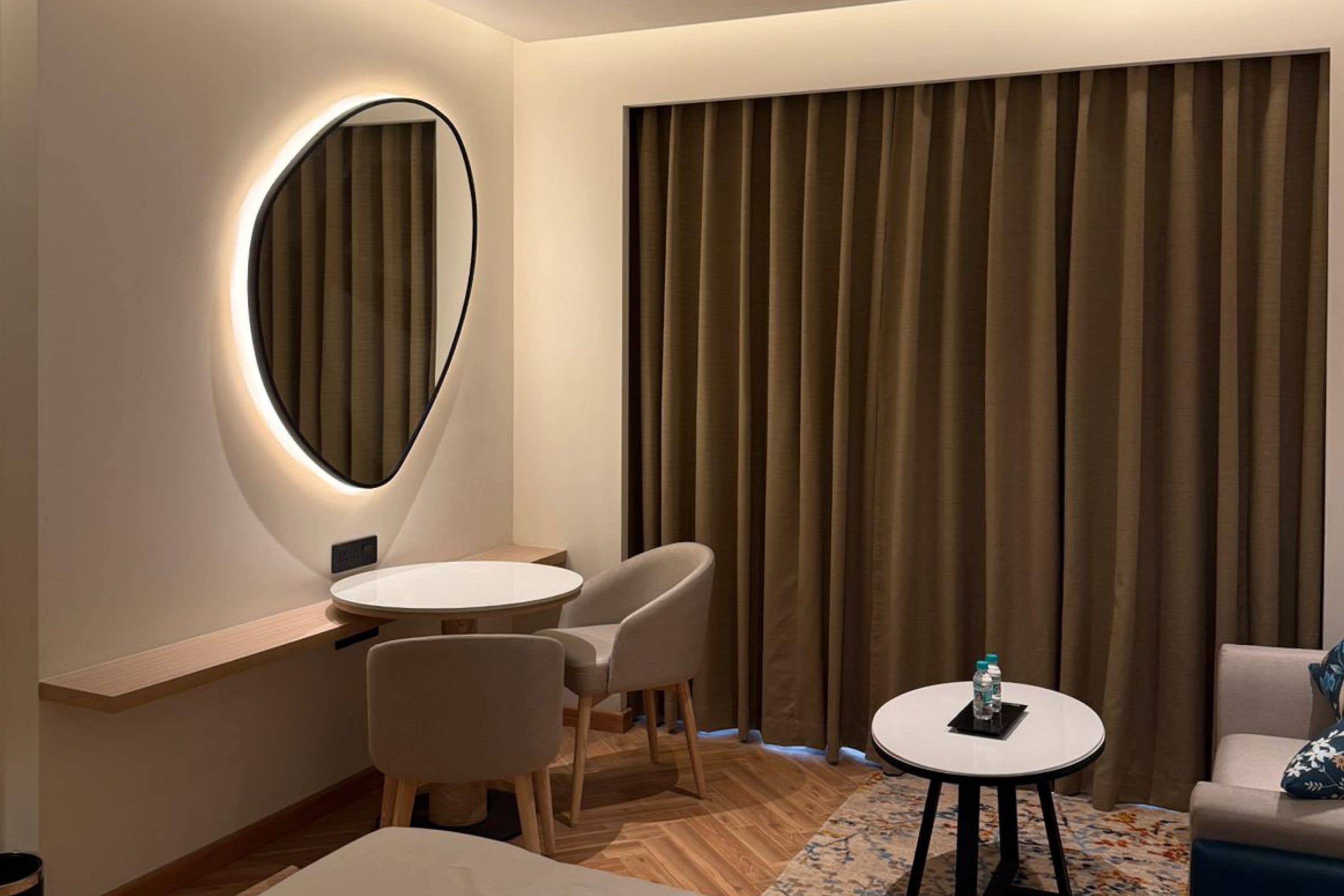 Habitación de hotel con una mesa redonda, dos sillas, una cama, una mesita con una botella y un ventanal grande con cortinas.