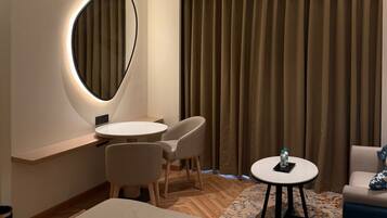 Habitación de hotel con una mesa redonda, dos sillas, una cama, una mesita con una botella y un ventanal grande con cortinas.