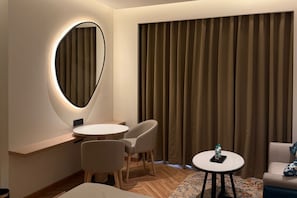 Habitación de hotel con una mesa redonda, dos sillas, una cama, una mesita con una botella y un ventanal grande con cortinas.
