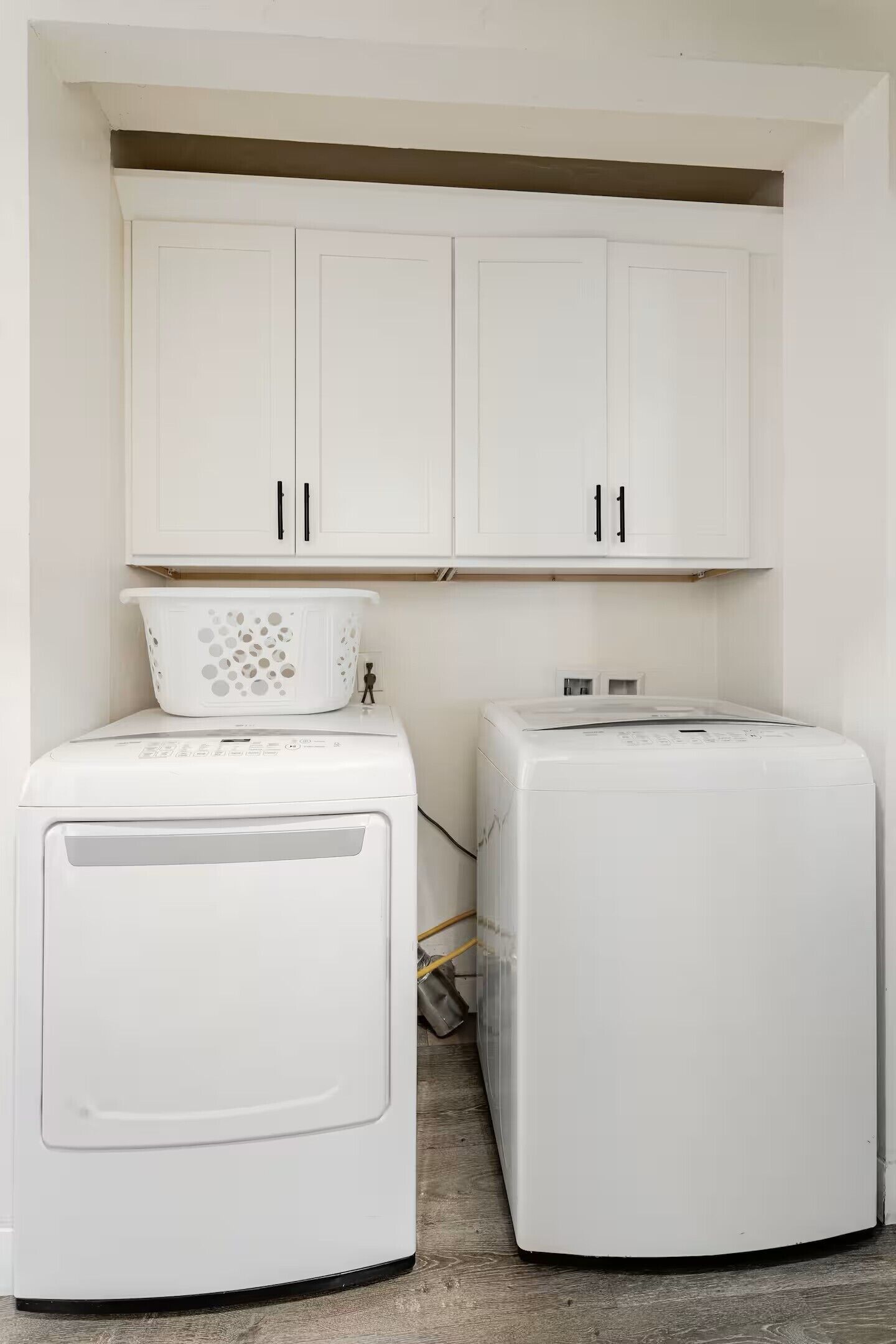 Een wasruimte met twee witte wasmachines, een wit kastje erboven en een witte wasmand aan de linkerkant.
