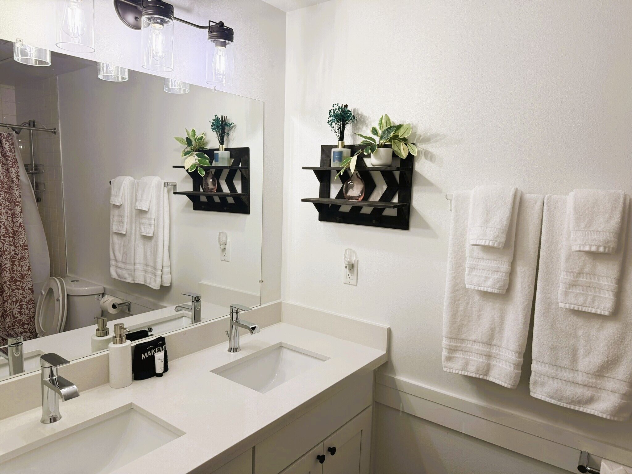 A modern bathroom with two sinks, a large mirror, towel racks, and decorative shelves with plants.