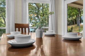 Una mesa de comedor con platos y tazones blancos, un florero con follaje verde y una silla de madera.