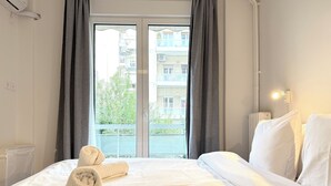 A hotel room with a bed, two towels, a window with curtains, and a view of buildings and greenery. - 1BR next to Pasalimani, Marina Zeas & Hospital (Pireas)