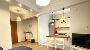 A modern living room with a sofa, a TV on a stand, and a kitchen area with a dining table and chairs. - 1BR next to Pasalimani, Marina Zeas & Hospital (Pireas)