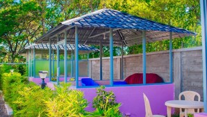 A covered outdoor seating area with a roof, surrounded by greenery and a purple wall.