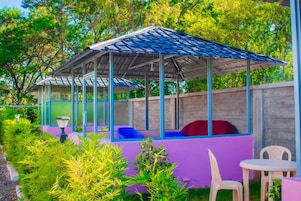A covered outdoor seating area with a roof, surrounded by greenery and a purple wall.