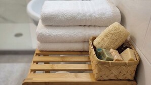 A wooden stool with a basket of toiletries, a stack of white towels, and toilet paper rolls in a bathroom.