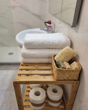 Ein hölzerner Hocker mit einem Tablett für Toilettenartikel, einem Stapel weißer Handtücher und Toilettenpapierrollen in einem Badezimmer.