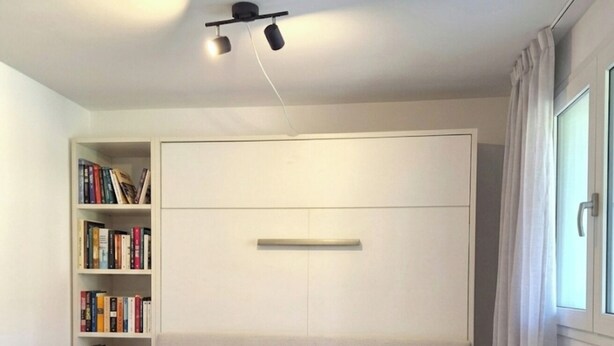 A modern hotel room with a large bed, a bookshelf, and a ceiling light fixture.
