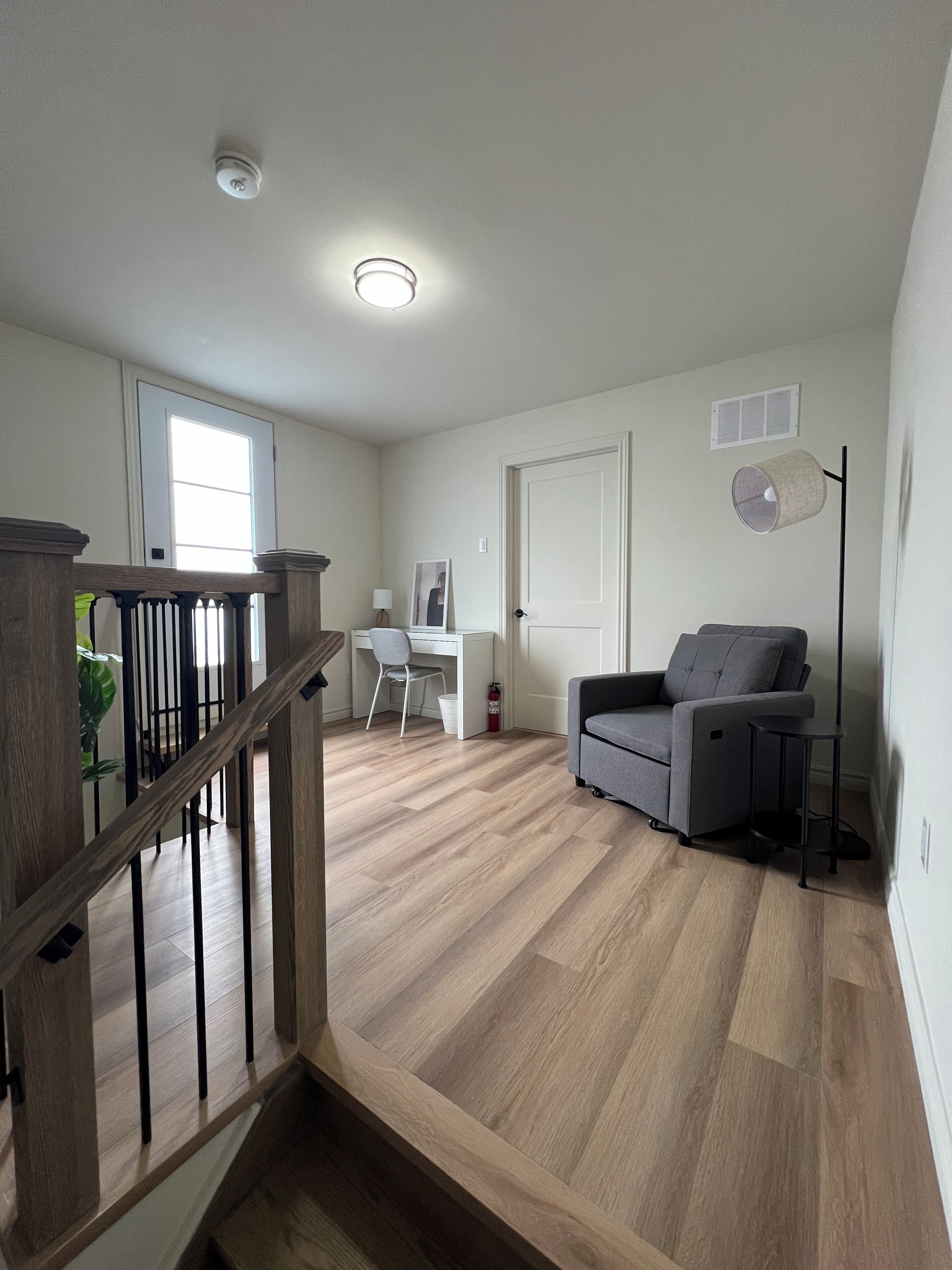 A spacious living area with a wooden staircase, a grey sofa, a white desk, and a ceiling light.