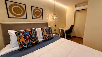 Una cama bien tendida con dos almohadas, un escritorio pequeño y un equipo de aire acondicionado en una habitación con una pared lisa y una lámpara colgante.