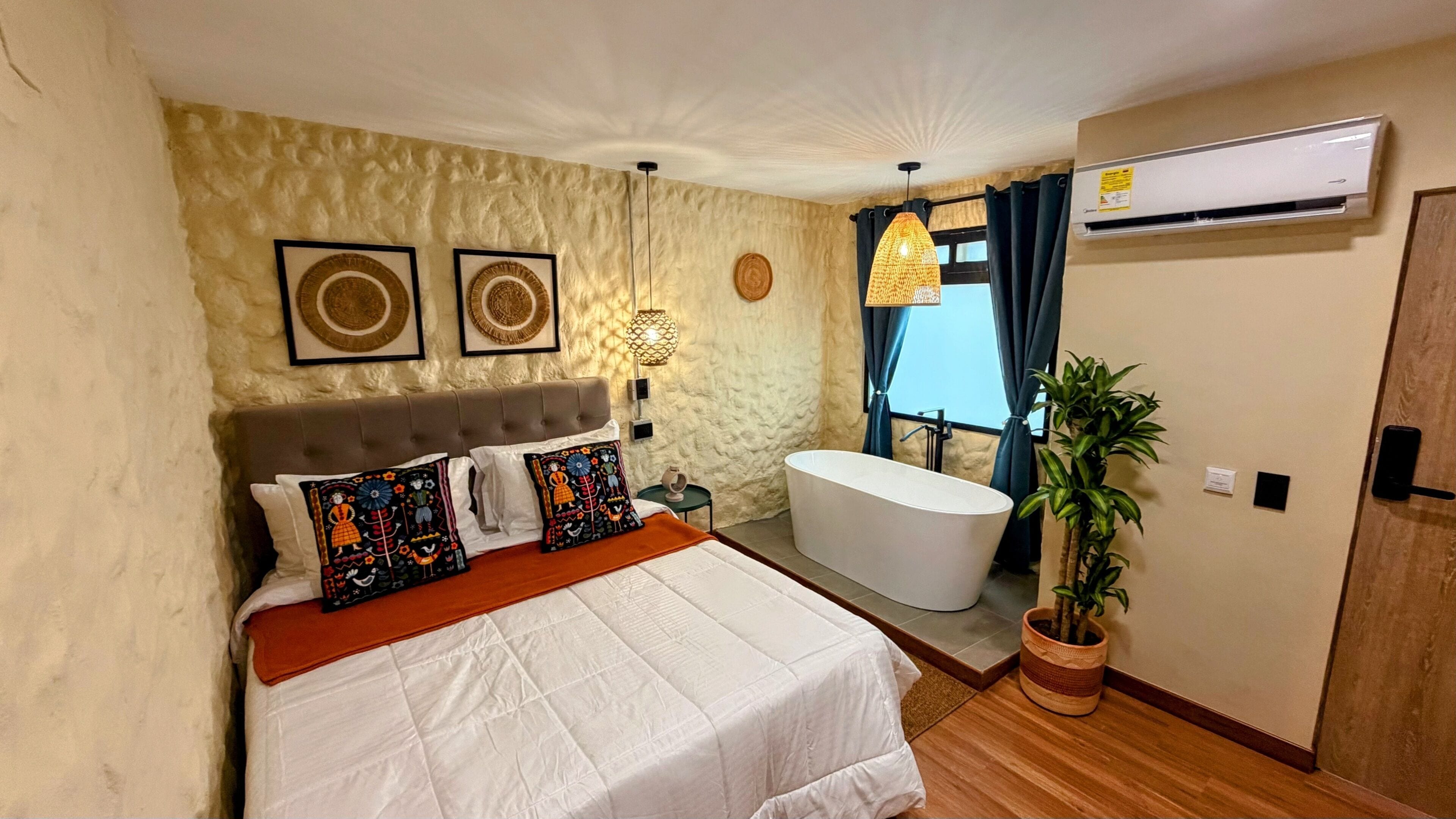 A bedroom with a bed, a bedside table, a wall-mounted air conditioner, a potted plant, and a bathroom with a bathtub.