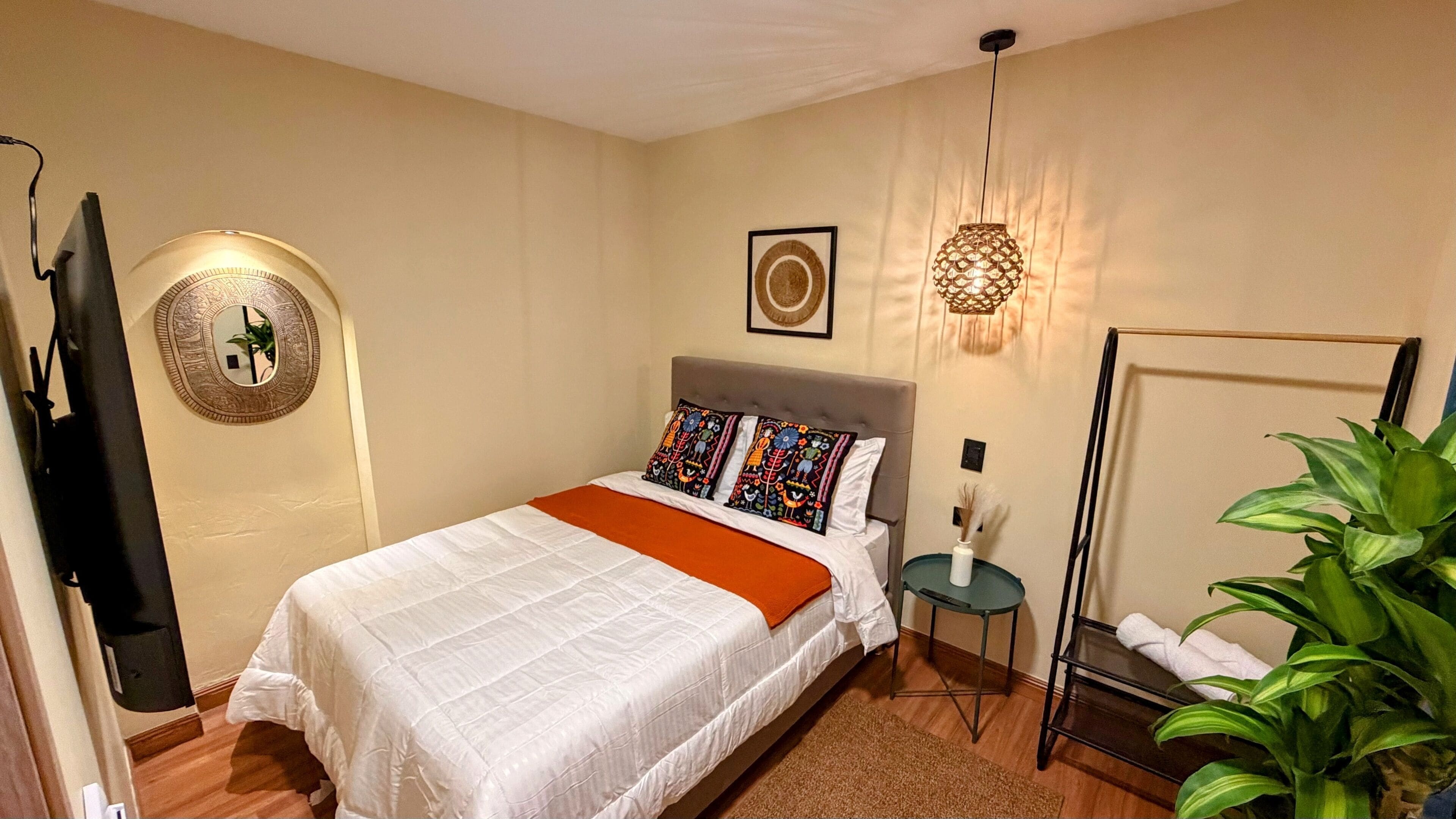 Una cama bien tendida con una colcha blanca y un plaid naranja, un cabecero tapizado en gris y un espejo fijado a la pared.