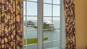 A view through a window with floral curtains, a glass balustrade, and a body of water with buildings in the distance. - Bayfront Efficiency with Pool and Gym (Ocean City)