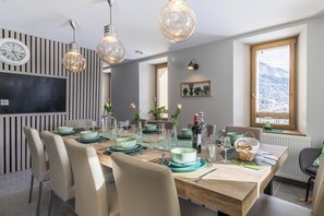 Een eetgedeelte met een houten tafel voor vier personen, omringd door stoelen. Er hangen hanglampen, een klok aan de muur en een raam met uitzicht op met sneeuw bedekte bergen.