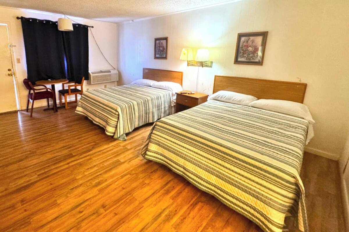 Et hotelværelse med to senge, et skrivebord, stole, en lampe og et vindue med gardiner.