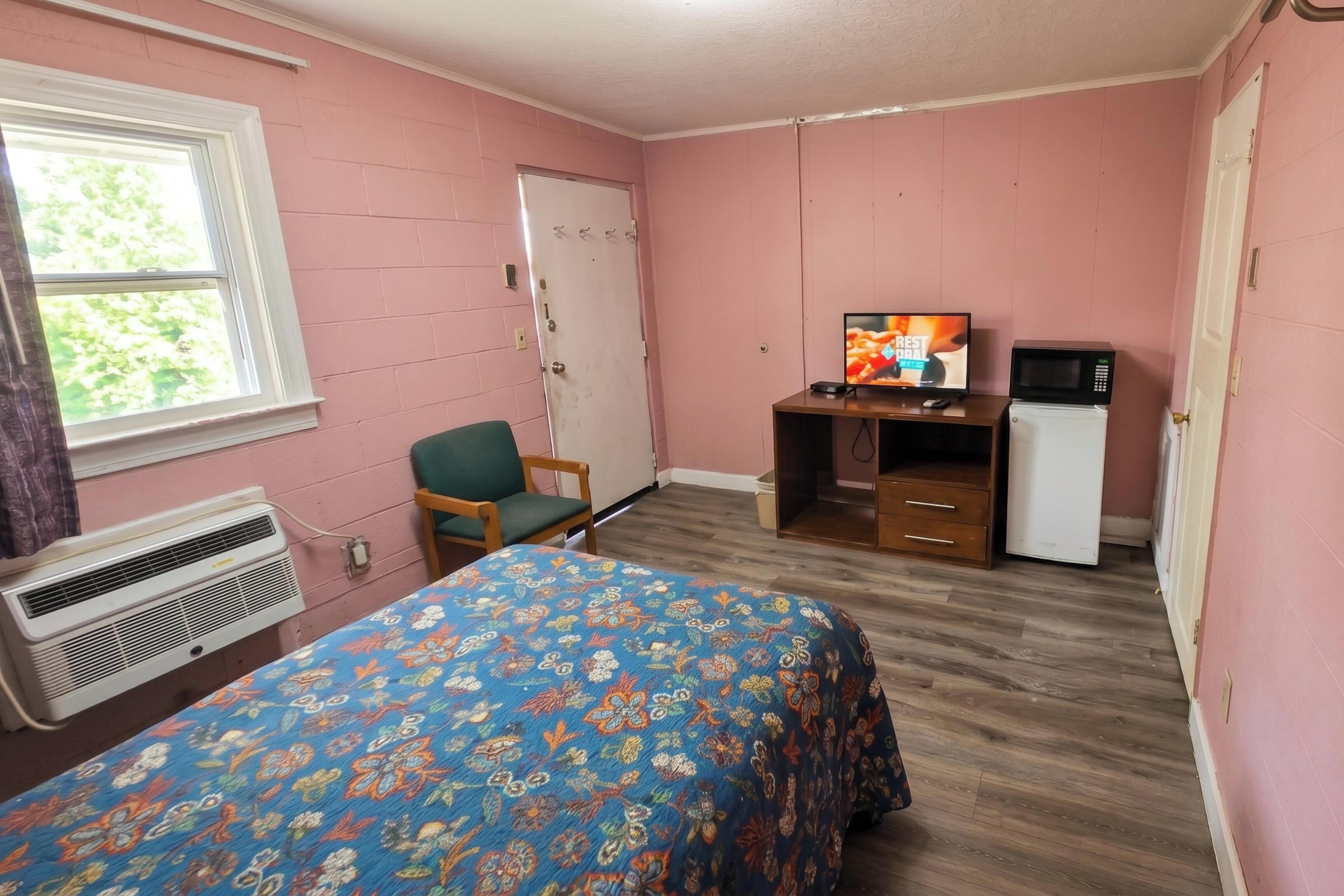 A room with pink walls, a bed with a floral bedspread, a desk with a television, a microwave, and a refrigerator.