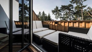Ett modernt utomhusområde med sittplatser, möbler i rotting och kuddar, en glasdörr som leder in, och ett trästaket med träd i bakgrunden.