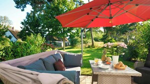 Un patio con giardino, con un ombrellone rosso, un divano in vimini e un tavolo apparecchiato per la colazione.