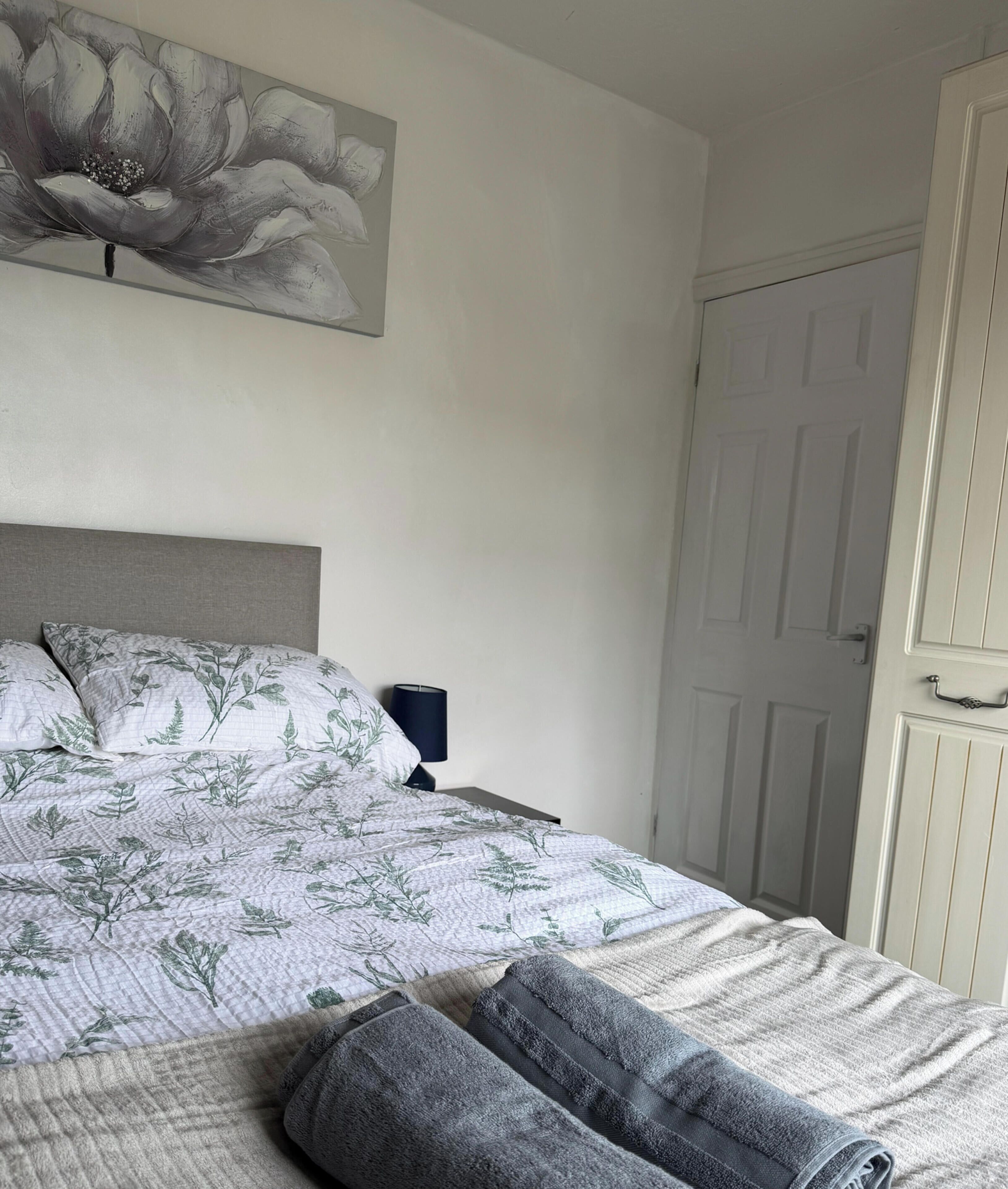 Un dormitorio con cama, mesita de noche, lámpara, puerta y un cuadro en la pared.