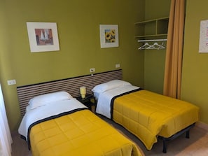 Una habitación con dos camas, una mesita de noche, una estantería con objetos colgando y una ventana con cortinas.