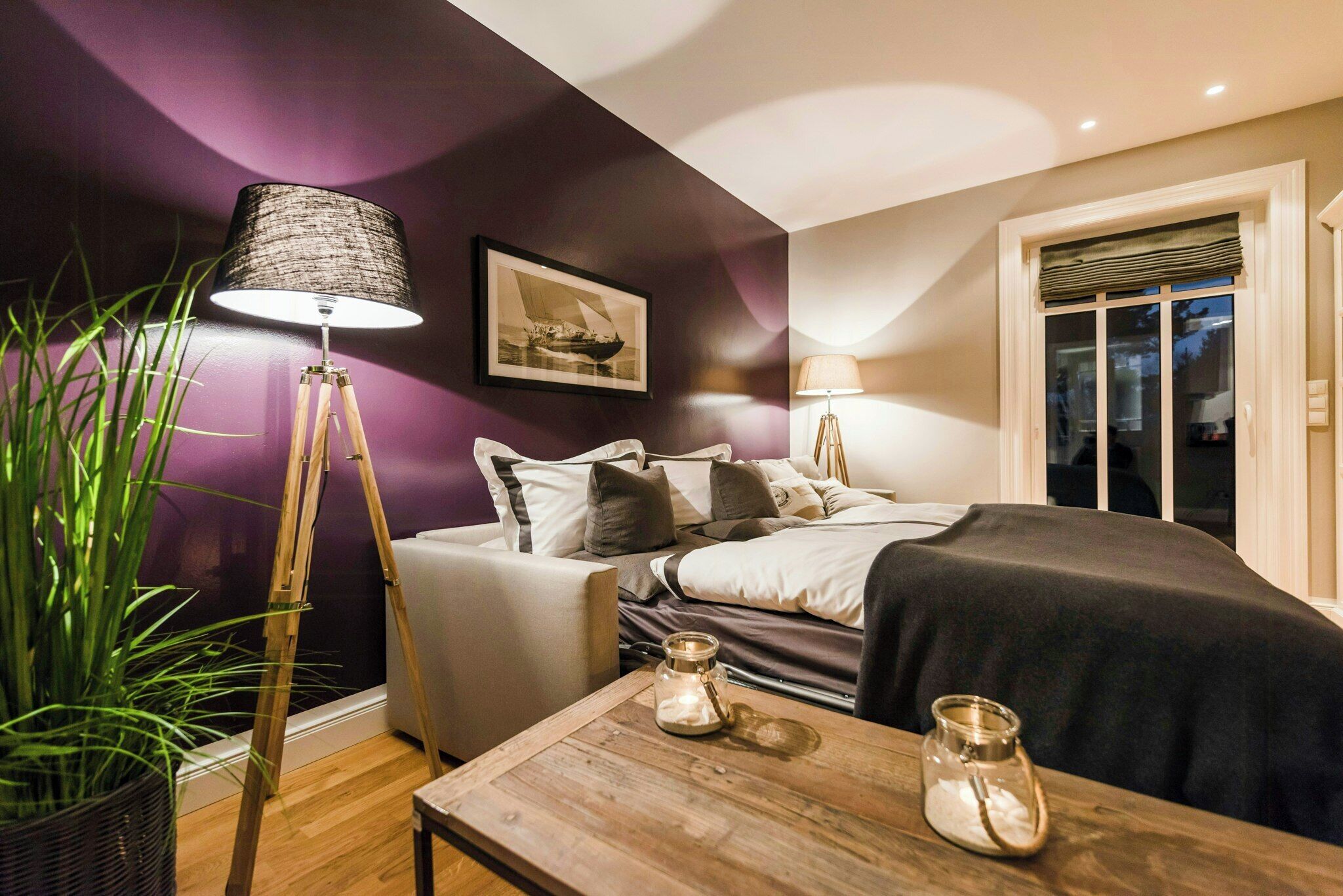 A bedroom with a bed, bedside lamps, a window, a potted plant, and a wooden table with candles.