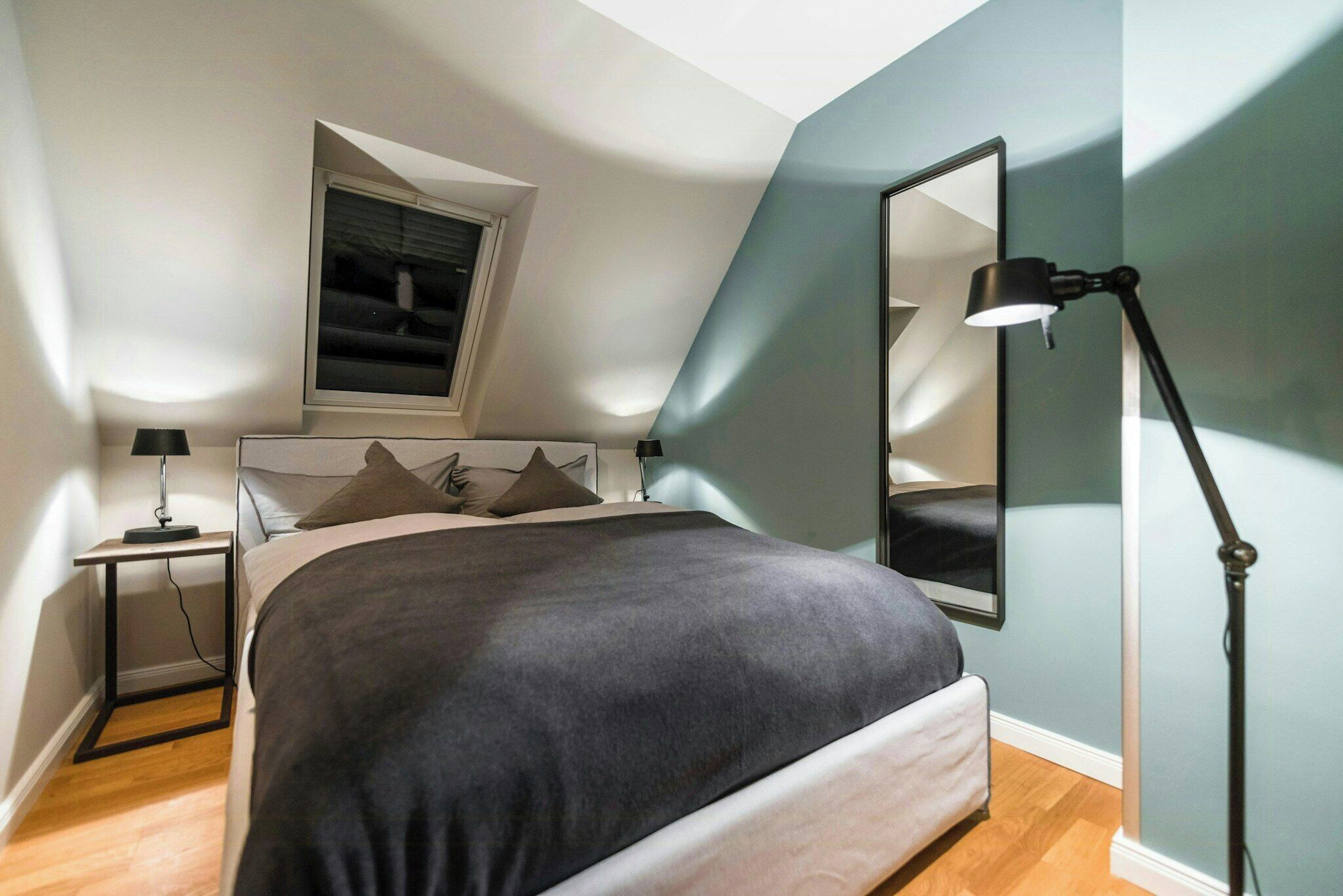 A bedroom with a bed, a nightstand, a desk, a lamp, a window, and a mirror.
