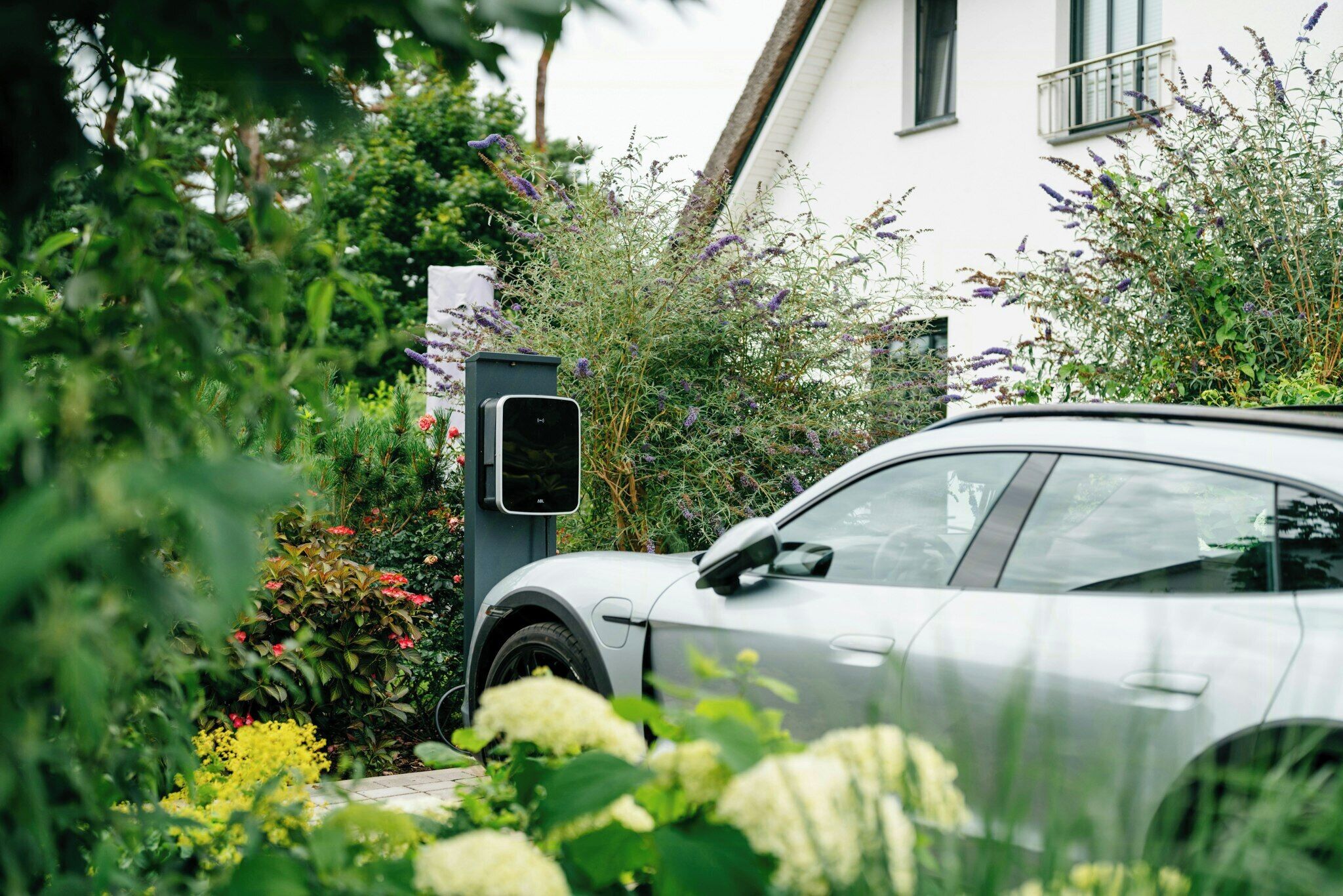 Une voiture est garée dans un jardin, à proximité d’une borne de recharge, et est entourée de différentes plantes. Un bâtiment se trouve en arrière-plan.